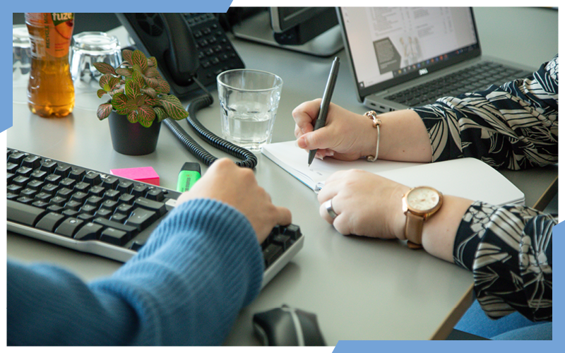 Das Bild zeigt eine Person an einer Tastatur, eine Person schreibt mit einem Kugelschreiber in einem Notizheft. Daneben steht ein Arbeitslaptop und Getränke. | © die media GmbH